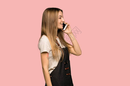 穿着围裙的年轻女青年以孤立的粉红背景与移动电话保持对话图片