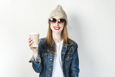 一个穿着牛仔夹克眼镜和帽子的年轻女孩拿着一杯白色背景的咖啡或茶图片