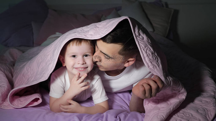 爸和他笑着的儿子在睡觉前躲在粉红色的毯子下黑暗图片