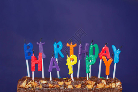 与花生盐焦糖和紫色背景上的彩色无光蜡烛一起的生日快乐布朗尼蛋糕图片