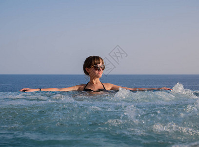 年轻的黑发女人在海空背景下在无穷的按摩浴图片