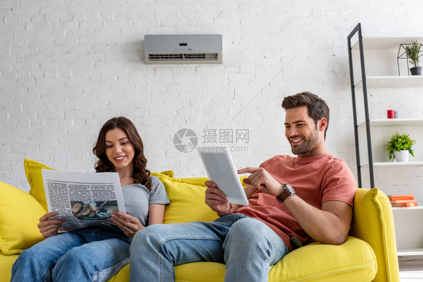 在家中空调机下坐在黄色沙发上时使用数字石板阅读报纸和英俊男子的美貌女图片