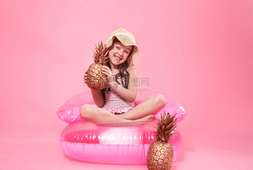 一个快乐的夏日小女孩穿着暑假帽子坐在一个充气椅上用金漆菠萝彩色背景夏天概念和创造力图片
