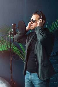 身穿太阳眼镜牛仔裤黑衬衫和外套的男歌手用耳机近距离在家庭工作室录制图片
