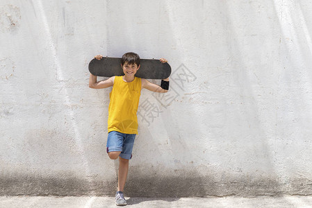 年轻笑的男孩靠在黄墙上手持滑板在背景图片