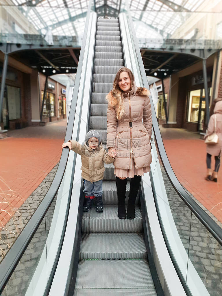 与美丽的年轻母亲在购物中心扶梯上一起微笑的幼图片