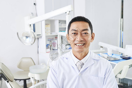 成功的成熟牙医在现代牙科诊所站立时用相机微笑的镜图片