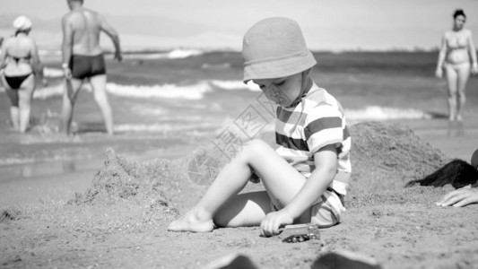 可爱的蹒跚学步的男孩戴着帽子坐在沙滩上玩具车的黑白照片孩子在暑假图片