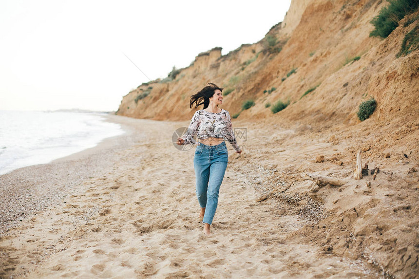 时尚的时髦女孩在海上的海滩上奔跑和微笑穿着牛仔裤和花卉衬衫的快乐波西米亚女人在热带岛屿的沙质悬崖上放松旅图片