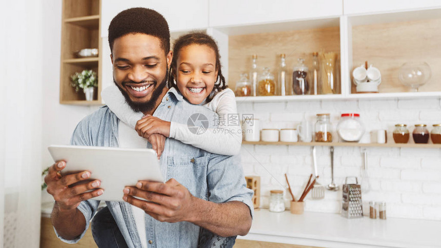 快乐的Afro孩子女孩拥抱她的父亲当他们一起看平板电脑在厨图片