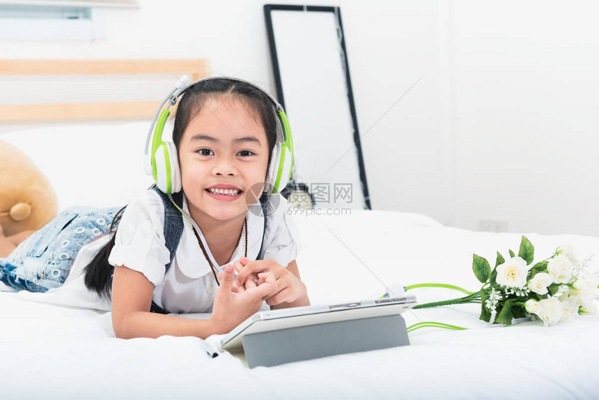 亚洲小女孩在家的床上用绿色耳机听平板电脑上的音乐通过倾听儿童和增强儿童的想图片