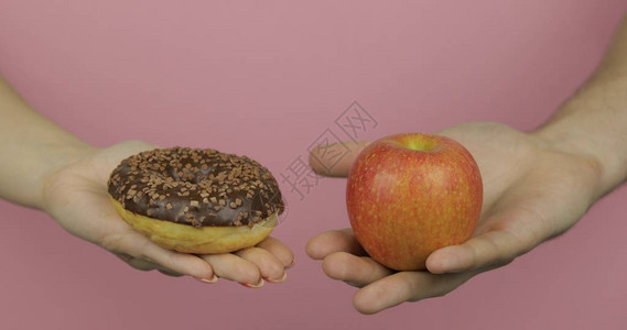 对苹果的选择甜圈开始健康饮食或垃圾食品粉红色背景中的手图片