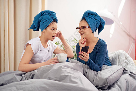 两个美丽的高加索女友头上戴毛巾坐在卧室的床上喝着咖啡和流言蜚语图片
