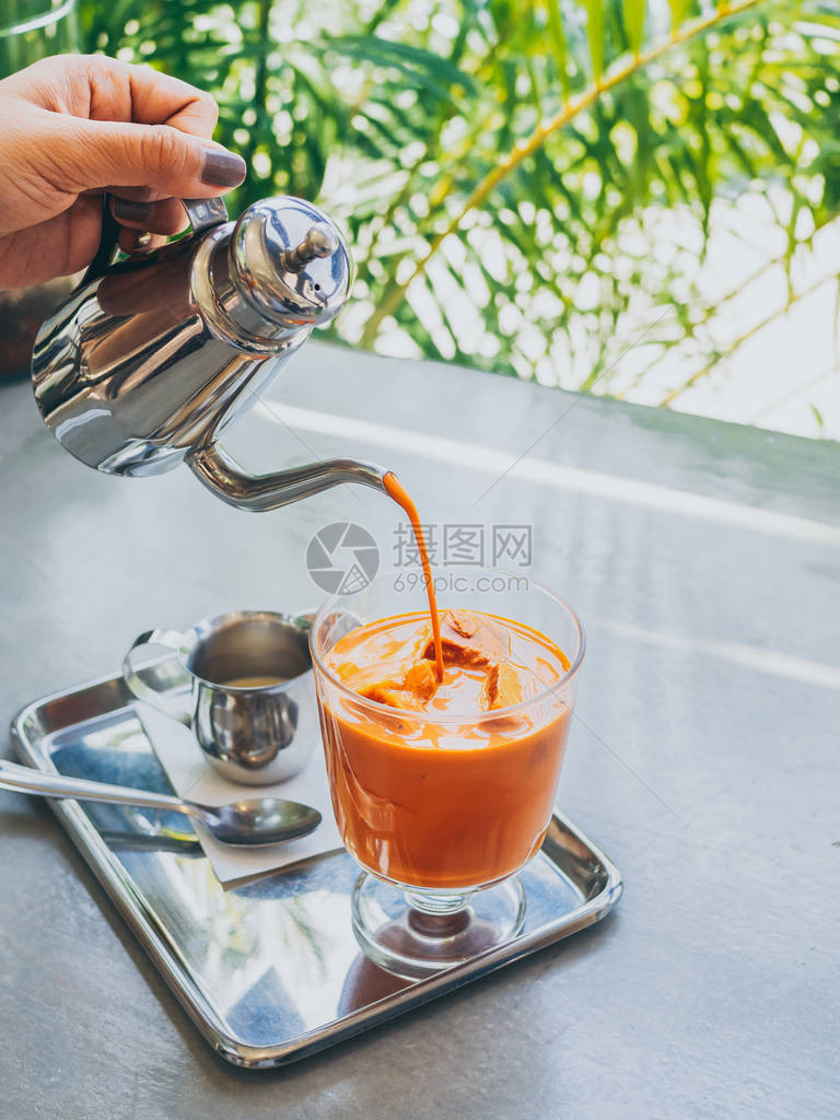 手工将不锈钢壶中的泰式奶茶倒在玻璃中的泰式茶冰块上图片