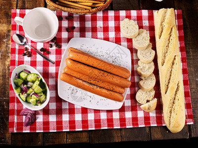 香肠部分与面包法式长棍面包放在格子桌布和农场风格的杯子上的桌子上图片