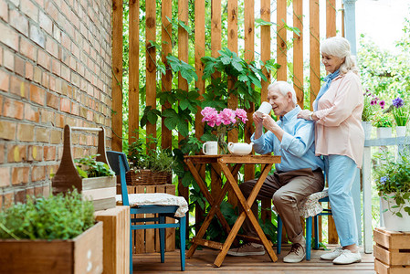 喜悦的年长妻子在丈夫旁边站着喝茶时有图片