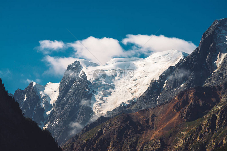 山脉顶部的低云和薄雾湛蓝的天空下的冰川阳光明媚的日子里的雪山峰巨大的岩石山脊与雪大气简约的高地图片