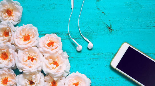 在旧漆木桌上放着新鲜的玫瑰智能手机和白色耳机图片