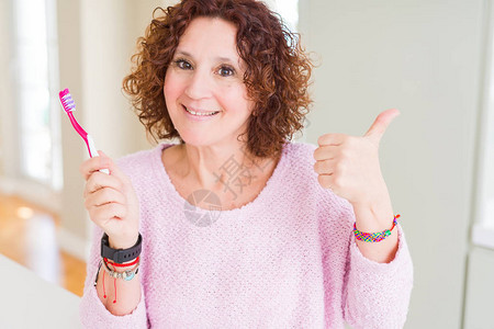 身持粉红牙刷的老年妇女在牙科诊所指着并用大拇指向侧面图片