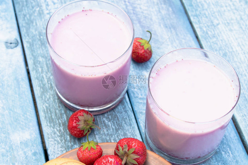 鸡尾酒加牛奶和浆果俱乐部在蓝板背景下的玻璃杯中牛奶和草莓的图片