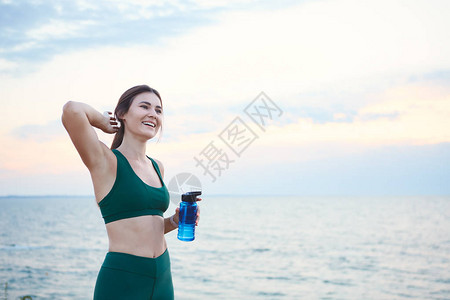 无线耳膜和智能手表的黑发年轻女人早上在海边日出饮用水时在海图片