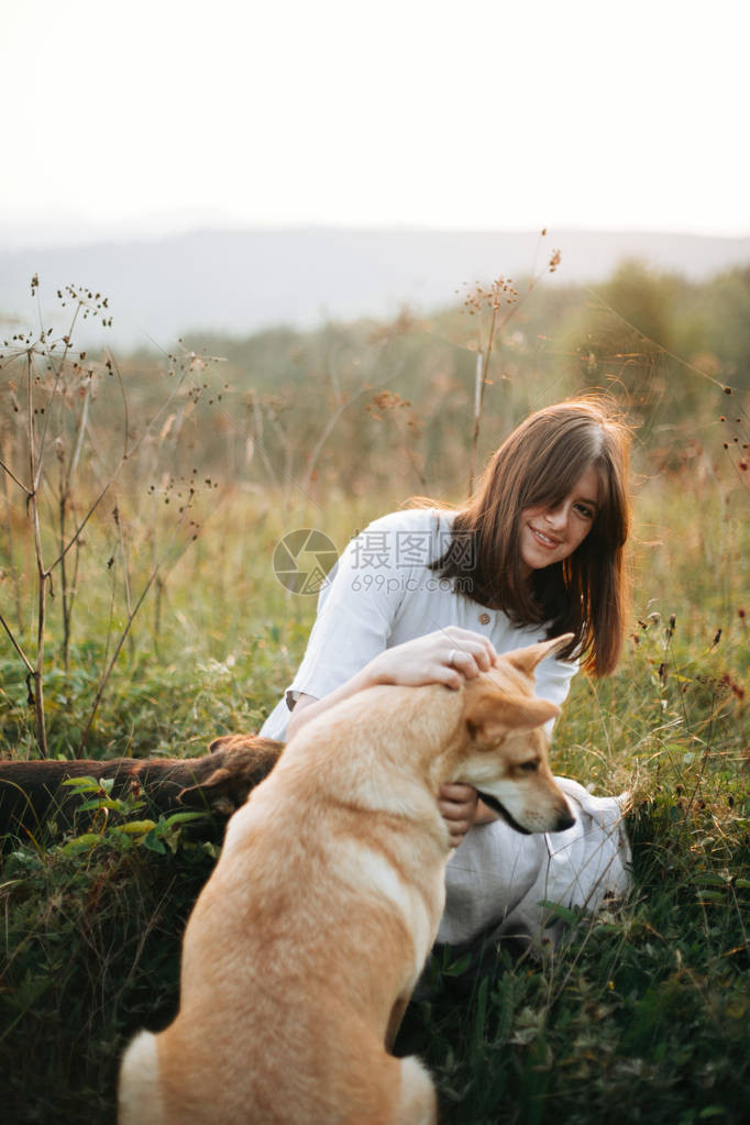 时尚的波西米亚女孩在日落时分在山上阳光明媚的草地上和她可爱的狗在草地和野花玩耍与宠物一起旅行爱抚她的金图片