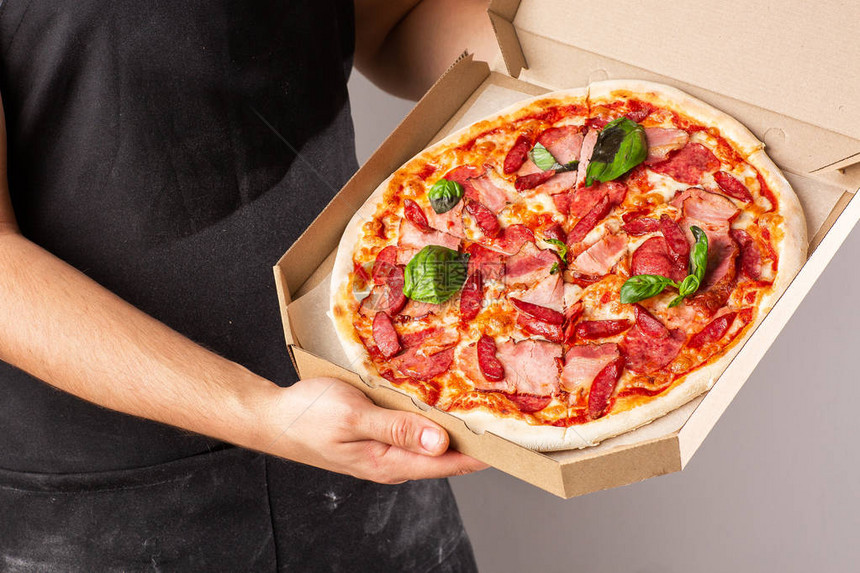 披萨和火腿和香肠放在黑围裙的年轻人手里的一个盒子里图片