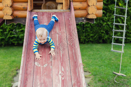可爱的白种人蹒跚学步的男孩在后院的生态友好型天然游乐场滑下木制滑梯图片