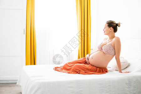 年轻的孕妇坐在床上抱着肚子微笑期待图片