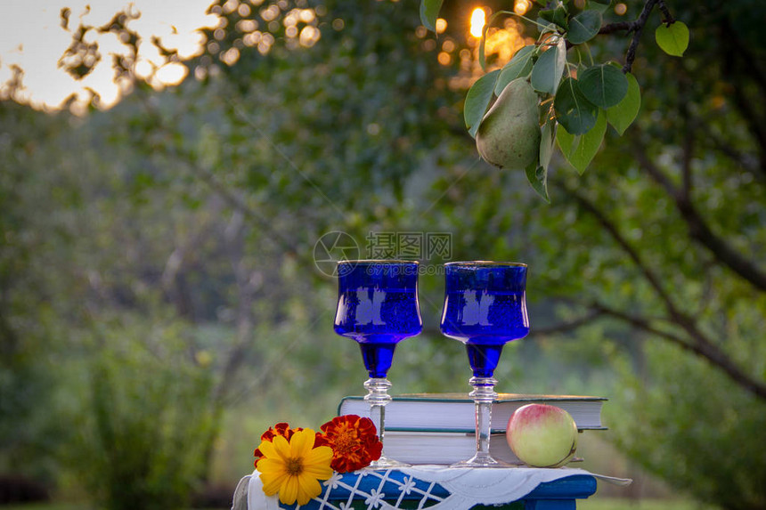 两个蓝色玻璃眼镜两本书一个苹果和一个用桌布装饰的凳子上的鲜花夏日花园图片