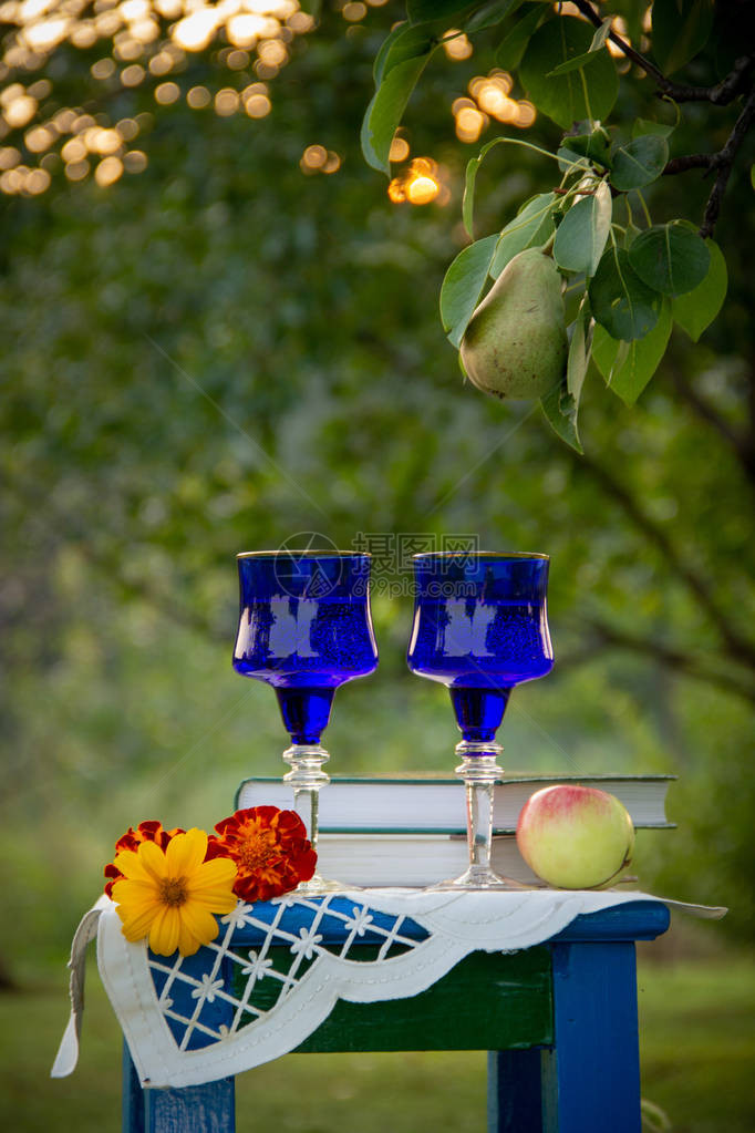 两个玻璃蓝色玻璃杯两本书一个苹果和一个用桌布装饰的凳子上的花图片