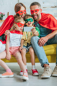 在超级英雄服装中快乐的家庭在家里用智能手机自拍时舌图片
