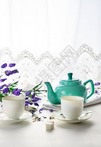 两个带绿茶的白茶杯茶色壶白色背景上的紫色花前视背景图片