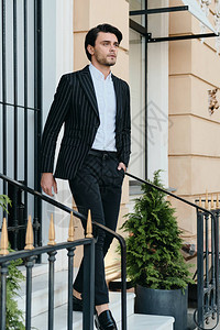 穿着白衬衫和经典夹克的黑发男子在街上的小楼梯上站着看一旁图片
