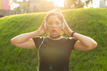 美丽感的年轻女子在阳光明媚的夜晚穿过公园时戴着耳机听音乐图片