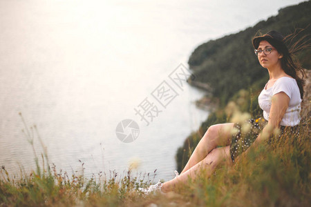 戴着帽子和风卷发的旅行时髦女孩坐在草丛中的岩山上图片