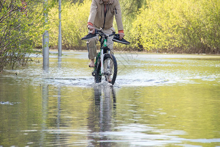 赤脚的骑自行车者试图克服春季洪水中的水随着河水冲破河岸图片