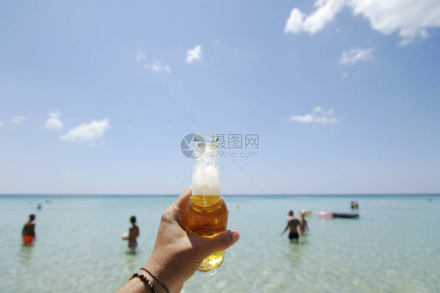 男手握着一瓶啤酒在阳光明媚的天空和清澈的海面上在背景中图片