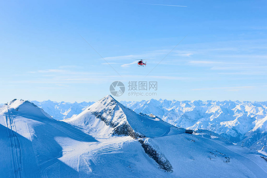 冬季在阿尔卑斯山的奥地利蒂罗尔州齐勒河谷的辛特图斯冰川滑雪胜地上空飞行红色直升机斩波器和高山与雪飞机和蓝天图片