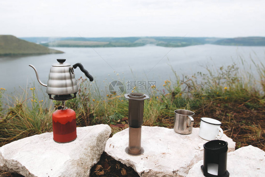 在湖背景的悬崖上煮沸的钢壶金属杯和图片