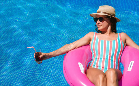在蓝泳池里戴太阳眼镜帽子和泳衣的年轻女子粉色浮标上的漂亮女孩在喝鸡尾图片