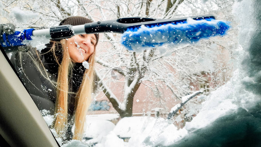 穿着帽子的年轻笑着女人试图清理她的汽车挡风玻璃和擦拭器在暴风雪过后用图片