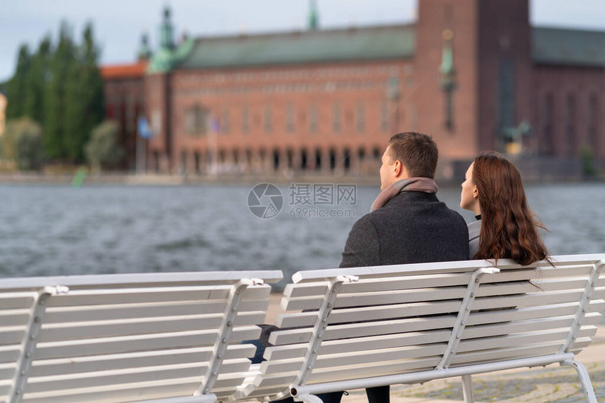 年轻夫妇在白色长椅上放松俯视着水和古老建筑在寒冷的冬天或秋天穿图片