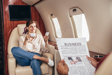 男朋友在私人飞机上看报纸和女朋友拿着杯子和智能手机图片