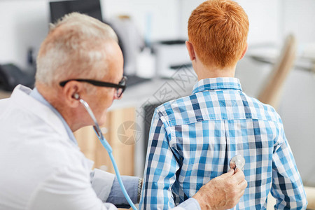 高级医生在儿童保健诊所咨询时用听诊器听可爱红发男孩心跳和呼吸的后图片