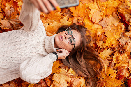 穿着时髦针织毛衣戴着时髦眼镜漂亮的年轻时髦女人躺在公园的橙叶上图片