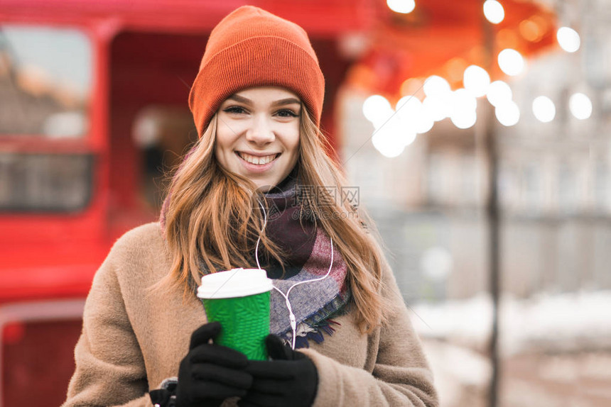 戴着耳机手里拿着一杯咖啡穿着暖和的衣服站在冬天户外明亮的背景下看着相机的微笑女孩的特写肖像一个女图片