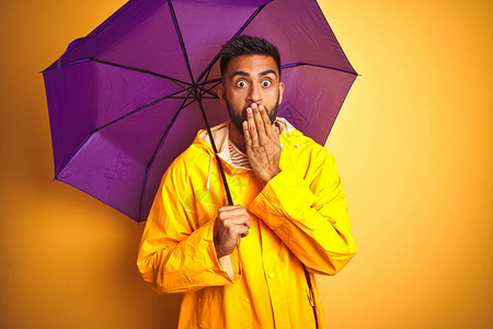 身穿雨衣和紫色雨伞的印度青年男子在孤立的黄色背景面罩上用手摇动嘴部图片