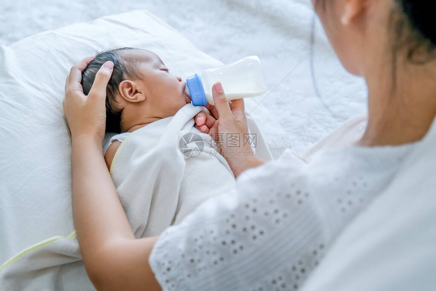 亚洲母亲用瓶子喂奶给白床上的小新生婴儿图片