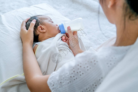 亚洲母亲用瓶子喂奶给白床上的小新生婴儿背景图片